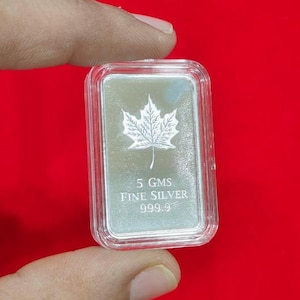 以下が含まれることがあります： 透明なプラスチックケースに入った、5グラムの長方形の銀の延べ棒。カエデの葉のデザインと「FINE SILVER 999.9」の文字が特徴です。背景は赤色です。