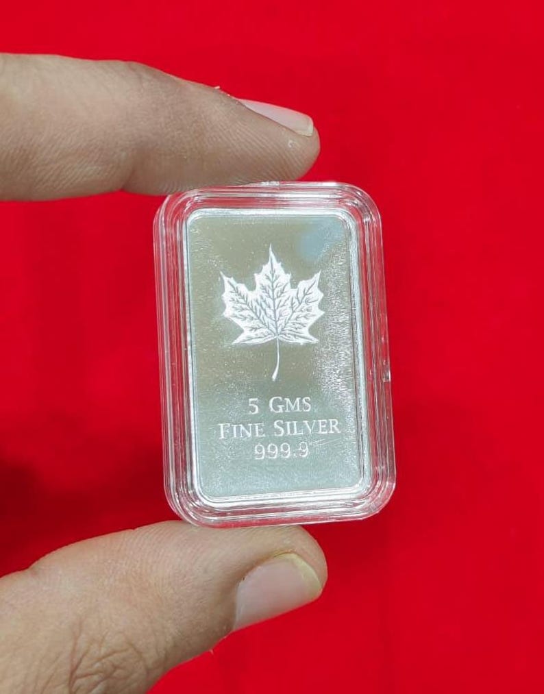 以下が含まれることがあります： 透明なプラスチックケースに入った、5グラムの長方形の銀の延べ棒。カエデの葉のデザインと「FINE SILVER 999.9」の文字が特徴です。背景は赤色です。