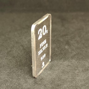 May include: A rectangular silver bar with the text "20g FINE SILVER 999" in white. The bar is slightly angled, revealing its thickness and a ridged edge. The background is a dark, solid color.