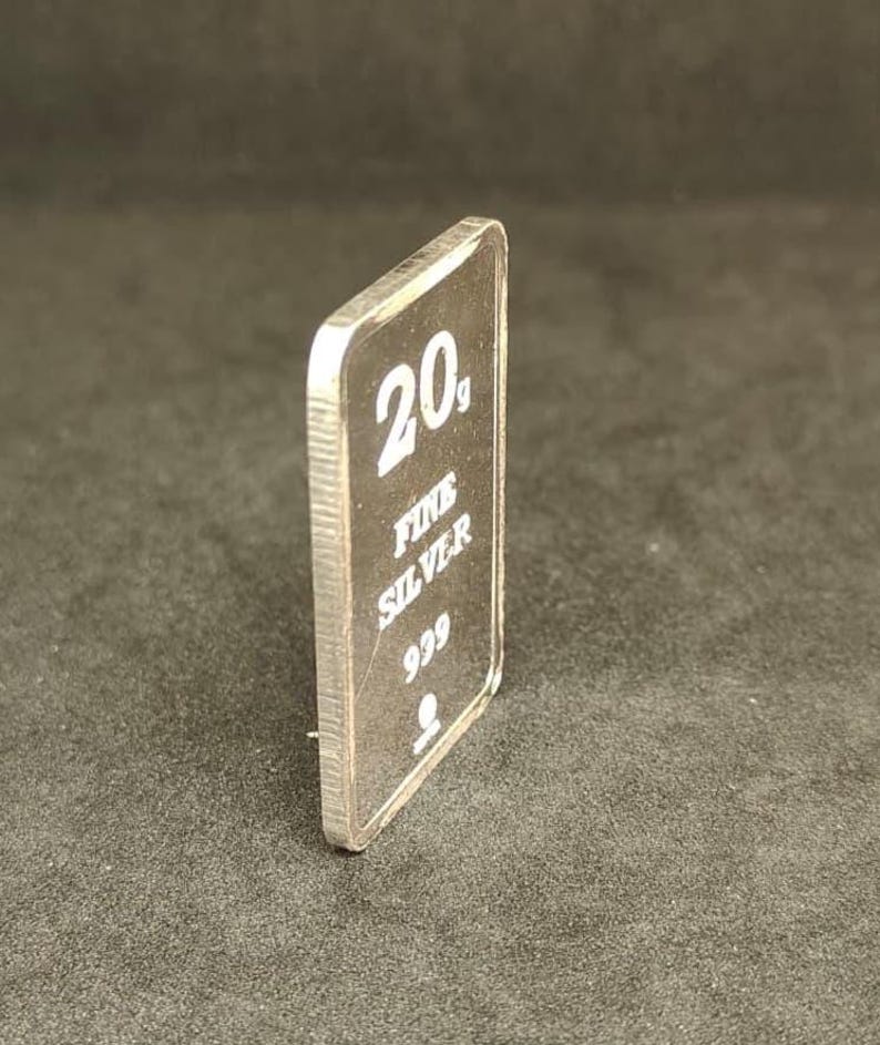 May include: A rectangular silver bar with the text "20g FINE SILVER 999" in white. The bar is slightly angled, revealing its thickness and a ridged edge. The background is a dark, solid color.