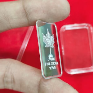 以下が含まれることがあります： カエデの葉のデザインが施された長方形の銀の延べ棒。「5 GMS FINE SILVER 999.9」と刻印されています。赤い背景を背景に、指で延べ棒を持っています。透明なプラスチックケースも確認できます。重量は5グラムです。
