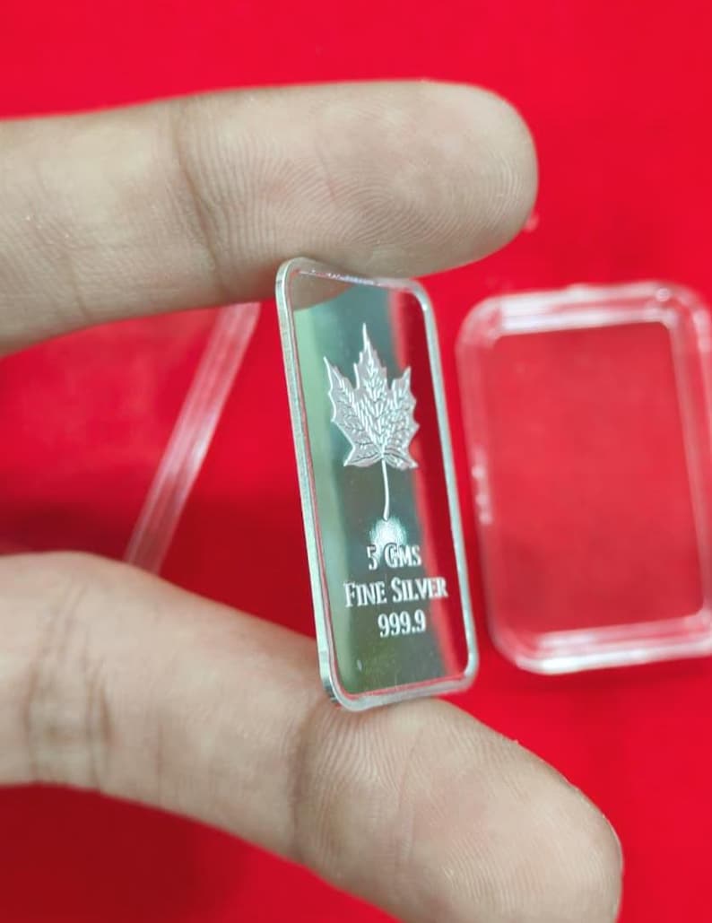 以下が含まれることがあります： カエデの葉のデザインが施された長方形の銀の延べ棒。「5 GMS FINE SILVER 999.9」と刻印されています。赤い背景を背景に、指で延べ棒を持っています。透明なプラスチックケースも確認できます。重量は5グラムです。