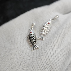 May include: Two silver fish pendants with red eye accents are displayed against a white background. The pendants have detailed scales and fins, and are designed with a loop at the top for a chain. The fish are approximately 2.5 cm in length.