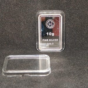 以下が含まれることがあります： 「10g FINE SILVER 999.9」と刻印された長方形の銀の延べ棒が、透明なプラスチックケースに入っています。 延べ棒には円形のロゴがあります。 ケースの蓋は分離されており、暗く質感のある表面に延べ棒の隣に置かれています。