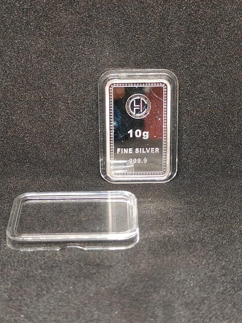 以下が含まれることがあります： 「10g FINE SILVER 999.9」と刻印された長方形の銀の延べ棒が、透明なプラスチックケースに入っています。 延べ棒には円形のロゴがあります。 ケースの蓋は分離されており、暗く質感のある表面に延べ棒の隣に置かれています。