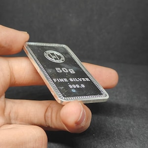 以下が含まれることがあります： 「50g FINE SILVER 999.9」の文字が入った長方形の銀の延べ棒。 延べ棒には装飾的な縁取りとロゴがあります。 延べ棒は手で持たれています。 重量は約50グラムです。