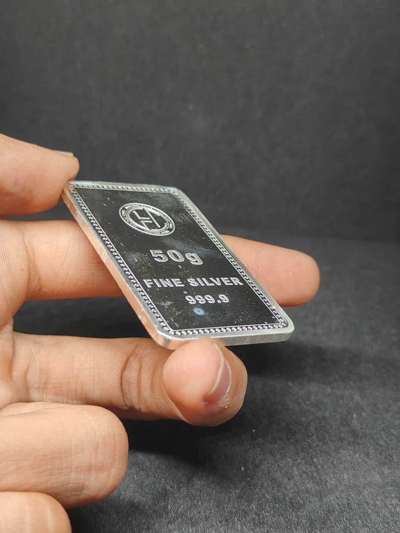 以下が含まれることがあります： 「50g FINE SILVER 999.9」の文字が入った長方形の銀の延べ棒。 延べ棒には装飾的な縁取りとロゴがあります。 延べ棒は手で持たれています。 重量は約50グラムです。