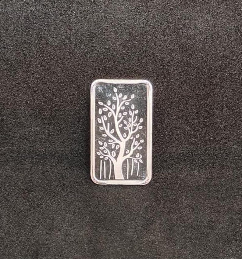 May include: A rectangular silver bar with a detailed tree design. The tree has a thick trunk, branches, and numerous leaves. The bar is set against a dark background.