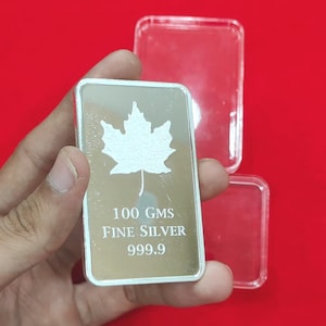 以下が含まれることがあります： カエデの葉のデザインが施された銀の延べ棒。バーには「100 GMS FINE SILVER 999.9」の刻印があります。赤い背景を背景に、手でバーが持たれています。透明なプラスチックケースがバーの後ろにあります。