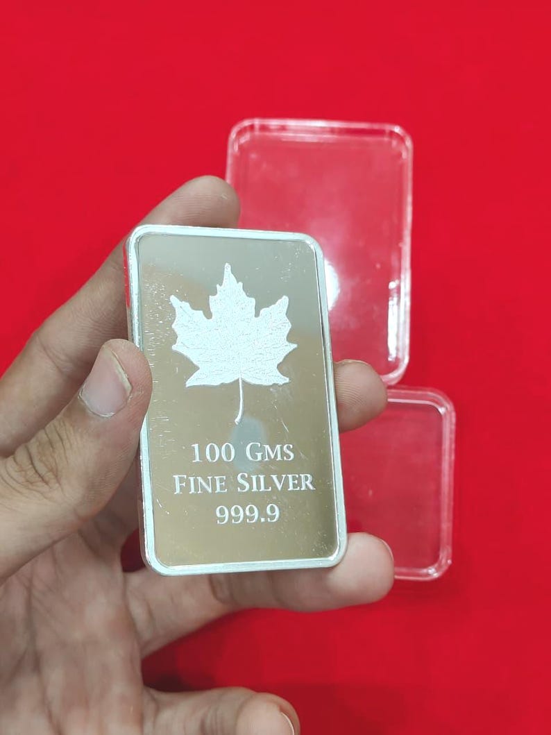 以下が含まれることがあります： カエデの葉のデザインが施された銀の延べ棒。バーには「100 GMS FINE SILVER 999.9」の刻印があります。赤い背景を背景に、手でバーが持たれています。透明なプラスチックケースがバーの後ろにあります。