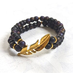 May include: A black beaded bracelet with a gold feather charm. The beads have a matte finish and are layered in a double strand.