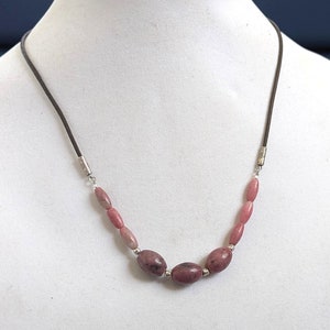 May include: A pink and brown stone necklace with silver accents on a brown leather cord. The necklace is displayed on a white mannequin bust.