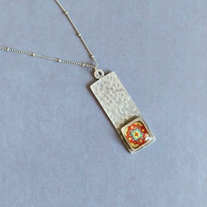 May include: A silver necklace with a rectangular pendant featuring a colorful tile design. The pendant has a hammered texture and the tile is set in a silver frame.