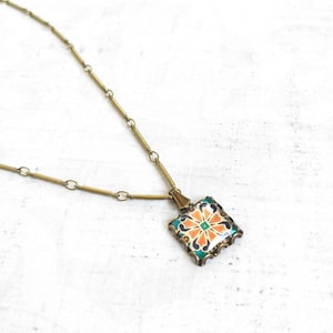 Puede incluir: Un collar de cadena dorada con un colgante cuadrado que presenta un diseño floral colorido. El colgante tiene un patrón verde, naranja y blanco.