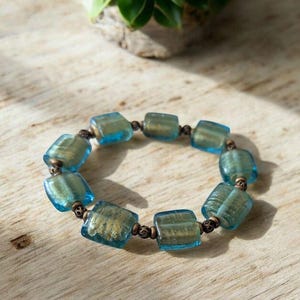 Może przedstawiać: Bransoletka z prostokątnymi, przezroczystymi niebieskimi koralikami ze złotymi plamkami. Koraliki połączone są małymi, ciemnymi metalowymi łącznikami. Biżuteria jest prezentowana na jasnej drewnianej powierzchni.