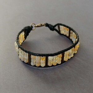 Puede incluir: Un brazalete de cuero negro con un cierre dorado y una fila de cuentas de color marrón claro y blanco.