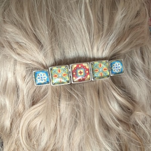 May include: A decorative hair clip with five square tiles. The tiles display colourful, intricate patterns in blue, yellow, red, and green. The clip is set against a backdrop of blonde hair.