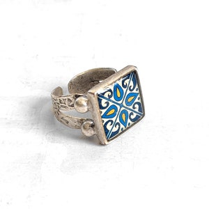 May include: A silver ring with a square blue, white, and yellow patterned tile design.