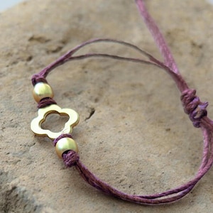 May include: A close-up of a handmade bracelet featuring a gold-colored clover charm and two gold beads. The bracelet is strung on a purple cord, knotted at each end. The background is a textured, neutral surface.