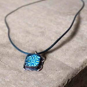 May include: A square pendant necklace with a blue and yellow floral design set in a copper-colored frame. The pendant hangs from a black cord, creating a simple yet elegant accessory.