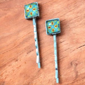 Op de afbeelding: Twee decoratieve haarspelden met vierkante, kleurrijke mozaïektoppen. Het mozaïekontwerp heeft gele, rode en blauwe patronen. De haarspelden hebben een zilverkleurige, zigzagvormige steel, tegen een houten achtergrond.