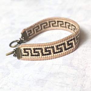 Op de afbeelding: Een beige kralen armband met een bruin Grieks sleutelpatroon en een bronzen sluiting.