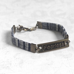 Puede incluir: Un brazalete de tela a rayas azul y blanco con un cierre de latón y una etiqueta rectangular de latón que dice "Fearless".