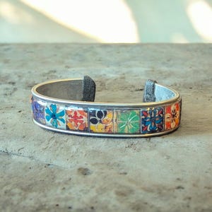 Op de afbeelding: Zilverkleurige manchetarmband met een mozaïekontwerp. De armband heeft een reeks kleurrijke, vierkante tegels met bloempatronen in blauw, oranje, groen en geel. De armband is aan de uiteinden open.