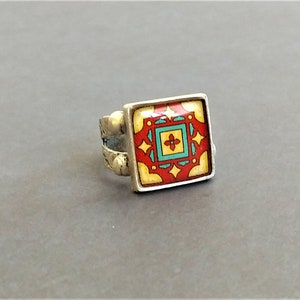 May include: A silver ring with a square-shaped red, yellow, and turquoise enamel design. The design features a geometric pattern with a central flower.