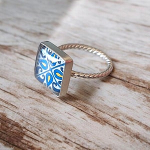 Puede incluir: Anillo de plata con una cara cuadrada con un patrón azul y blanco. La banda del anillo tiene un diseño de cuerda trenzada. El anillo está sobre una superficie de madera.