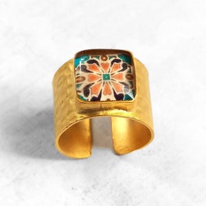 May include: A gold ring with a square, colorful tile design featuring a pink flower. The ring is made of hammered metal and has a wide band.