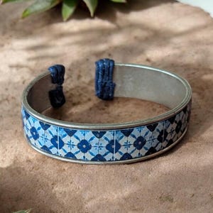 Puede incluir: Brazalete de puño plateado con un diseño floral azul y blanco. El brazalete tiene un diseño rectangular con un patrón repetido de flores azules sobre un fondo blanco. Los extremos están conectados con tela azul oscuro.