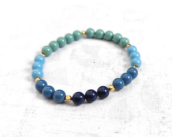 May include: A stretch bracelet featuring a gradient of smooth, round beads transitioning from light teal to medium blue and dark navy blue. Small, metallic gold-toned spacer beads are interspersed between the coloured spheres throughout the design.