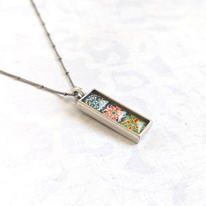 May include: A silver-toned necklace featuring a rectangular pendant with three colorful, patterned tiles. The tiles display intricate designs in shades of blue, red, yellow, and green. The necklace has a delicate chain with small, evenly spaced beads.