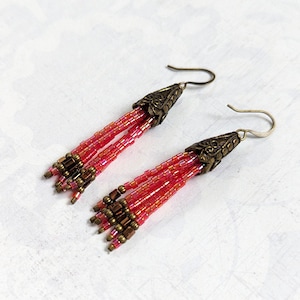 May include: A pair of long, beaded earrings in a pink and gold colour scheme. The earrings feature a tassel-like design with multiple strands of beads and a decorative, ornate, gold-toned metal top.