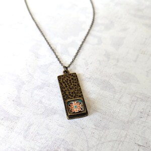 May include: A bronze necklace with a rectangular pendant featuring a colorful floral tile design.