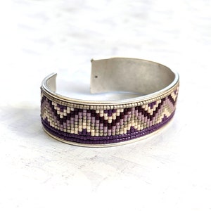 May include: A silver cuff bracelet with a purple, white, and beige beaded design. The beads are arranged in a geometric pattern.