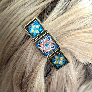 Puede incluir: Una pinza para el cabello con tres azulejos cuadrados engastados en un marco de metal color bronce. Los dos azulejos exteriores muestran un patrón floral azul y amarillo, mientras que el central presenta un diseño floral geométrico en naranja, blanco y azul.