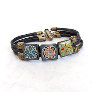 May include: A black leather bracelet with three square tiles featuring colorful geometric patterns. The tiles are set in gold-toned metal frames and are accented with gold-toned beads.
