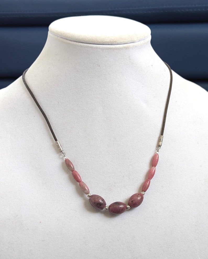 May include: A pink and brown stone necklace with silver accents on a brown leather cord. The necklace is displayed on a white mannequin bust.