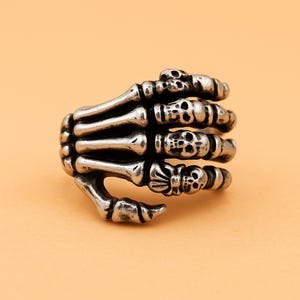Puede incluir: Un anillo plateado con forma de mano de esqueleto. El anillo presenta dedos esqueléticos detallados y pequeños detalles de calaveras en los nudillos. El anillo está sobre un fondo naranja claro.