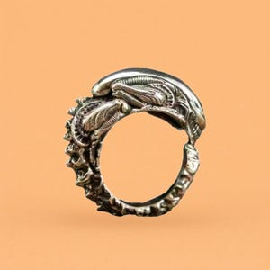May include: A silver-colored ring featuring a detailed design of an alien creature. The ring has a textured band and a prominent alien head and body design. The ring is set against a solid orange background.