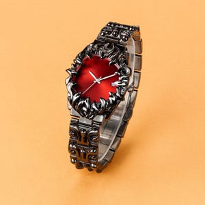 Pode incluir: Um relógio de pulso preto e vermelho com um design único. O relógio apresenta um mostrador vermelho escuro e uma moldura e pulseira pretas e ornamentadas. Os ponteiros do relógio são brancos. O relógio é exibido em um fundo laranja claro.