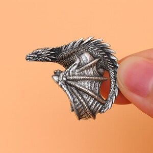 Könnte beinhalten: Ein detaillierter, silberfarbener Drachenanhänger. Der Drache ist mit aufwendigen Schuppen, Flügeln und einem eingerollten Schwanz dargestellt. Der Anhänger wird von Fingern gehalten, was die feinen Details des Designs zeigt. Ein Fantasy-Accessoire.