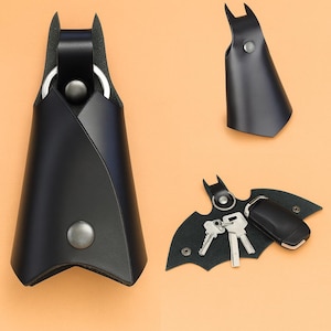 May include: Black leather key holder in a bat-wing design. The key holder has a metal ring and a snap closure. The image shows the key holder with keys attached, and a separate bat-wing shaped piece.
