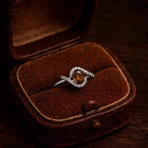 May include: A silver ring featuring an oval, amber-hued gemstone, encircled by small, clear stones. The ring is presented in an open, brown, velvet-lined jewellery box. The box is made of brown leather and rests on a dark wooden surface.