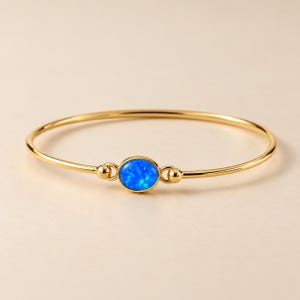 May include: A gold bangle bracelet featuring a central oval-shaped blue opal stone. The thin, polished gold band connects to the stone setting with small, rounded metal accents on each side, creating a sleek and continuous circular design.