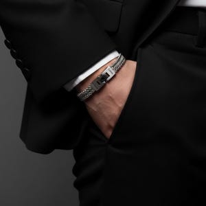 Op de afbeelding: Een zilveren gevlochten armband met een rechthoekige sluiting wordt om een pols gedragen. De armband is gemaakt van verweven metalen strengen. De sluiting heeft de merknaam gegraveerd. De persoon draagt een zwart pak met een wit overhemd.