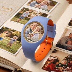 May include: A blue and orange wristwatch with a photo inside the watch face, resting on an open photo album. The watch has a blue band and an orange strap. Family photos surround the watch.