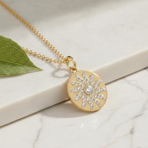 Puede incluir: Un collar con colgante dorado con un diseño de sol. El colgante circular presenta una piedra preciosa central transparente y acentos de cristal radiantes. La cadena del collar también es dorada.